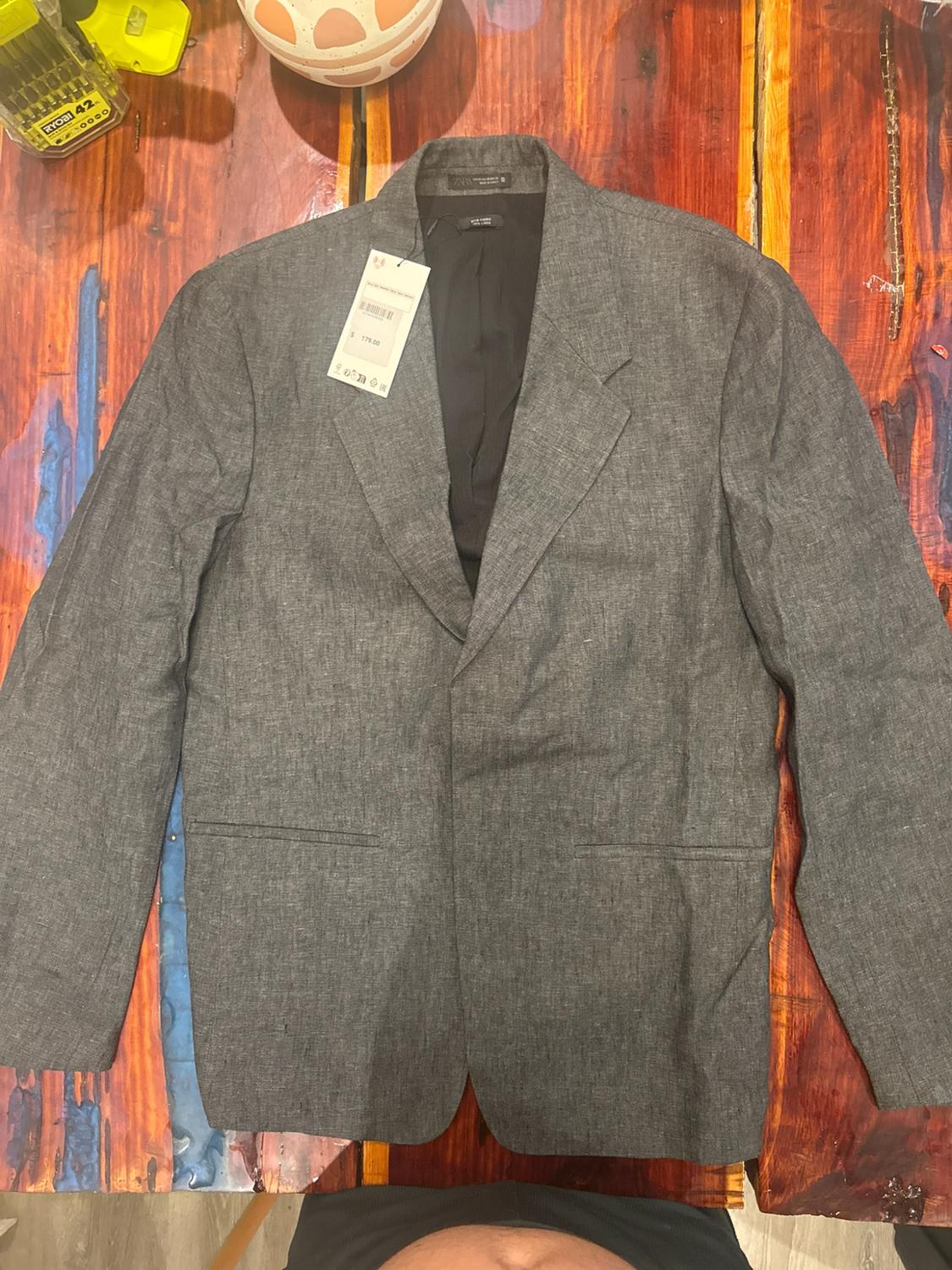 Grey blazer with label on a colourful rustic wooden table. Texts: RYOBI 42, $ 179.00, ZARA.