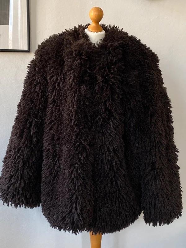 Dark brown fur coat on a wooden mannequin, hanging on a white wall with a framed picture in the background.