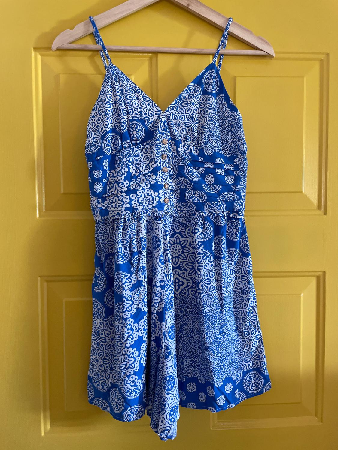 Blue jumpsuit with white print hanging on a wooden hanger over a yellow door.