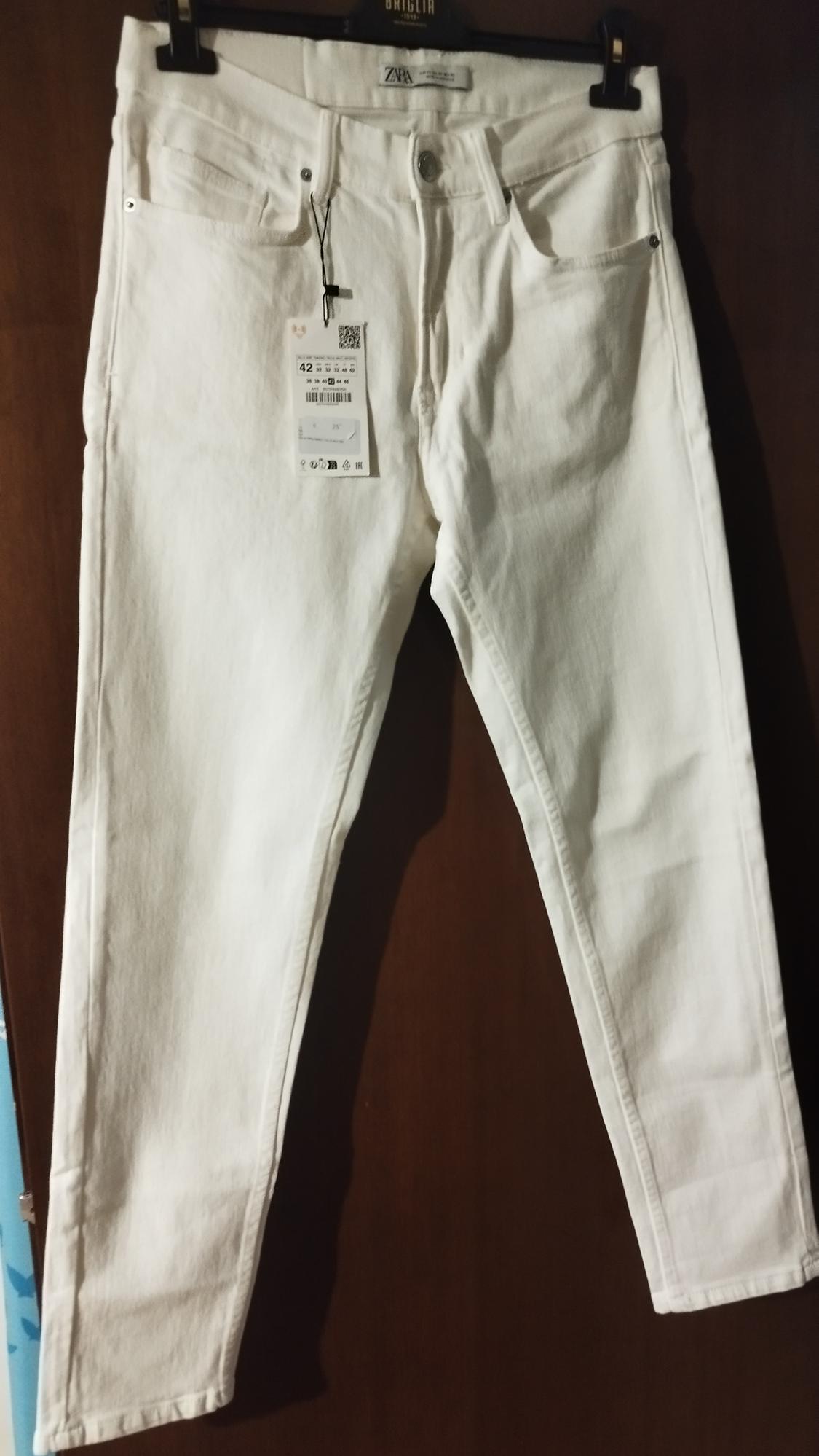 White trousers hanging on a black hanger with visible label. Dark wood background.