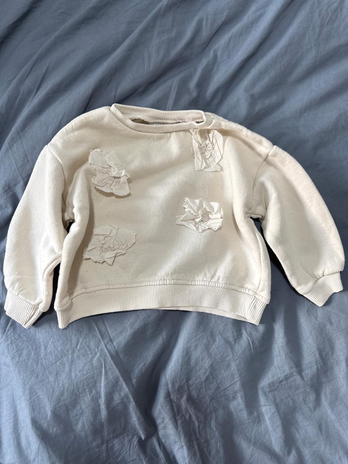 White sweatshirt with crinkled fabric details on a bed of grey sheets. Casual and relaxed style.