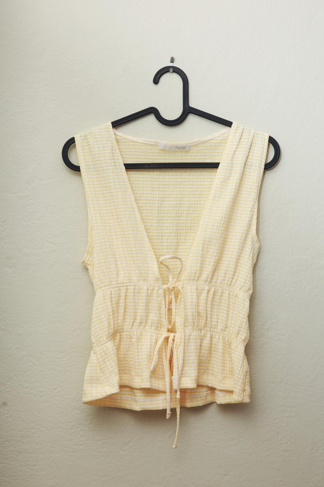 Yellow and white checkered sleeveless top on a black hanger against a light-colored wall. V-neckline and front tie closure.