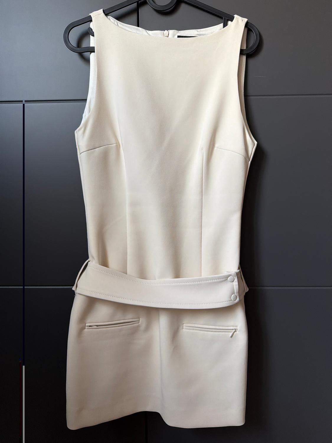Cream sleeveless dress hanging on a black hanger, with matching belt and front pockets, on a dark background.