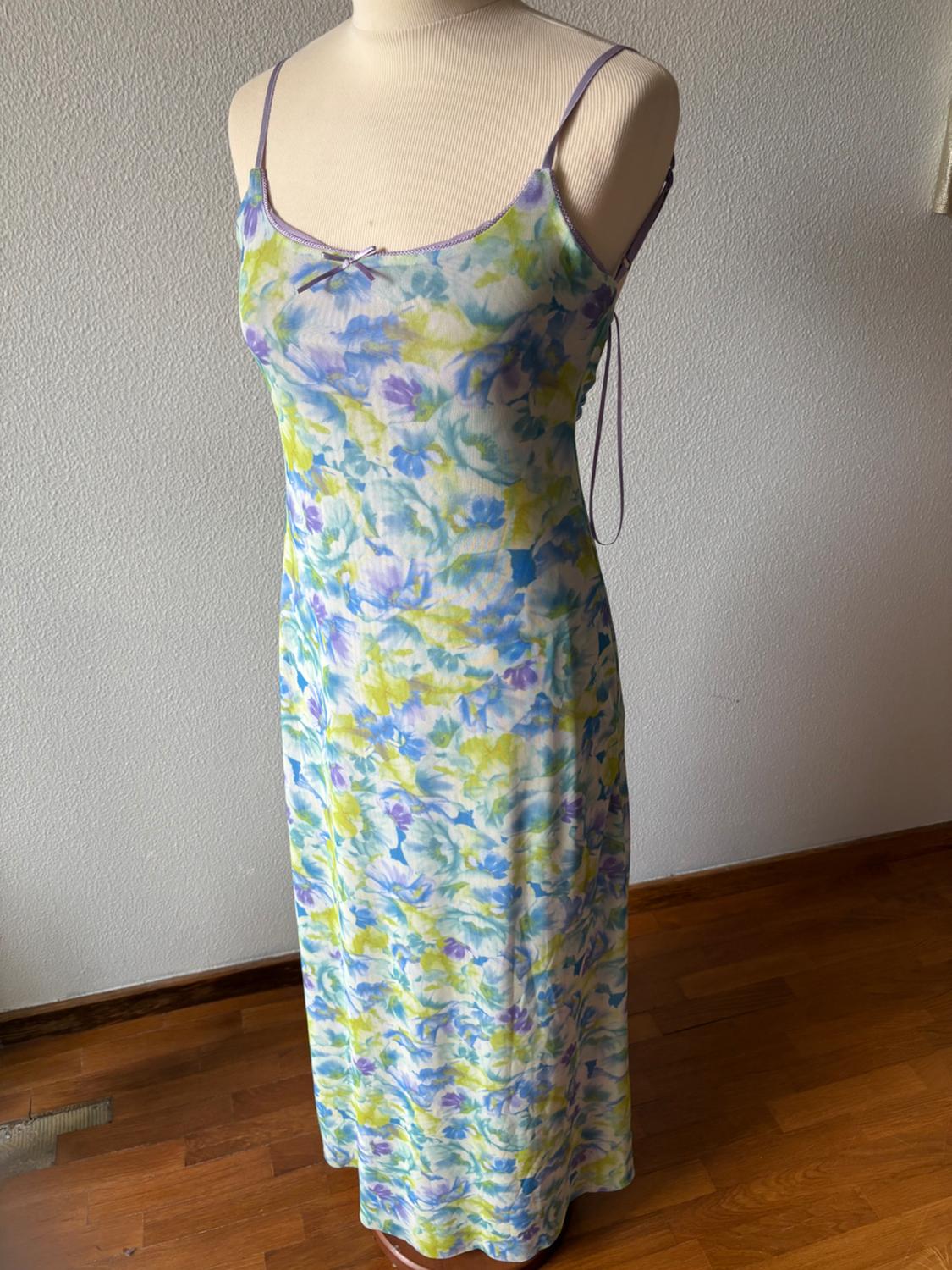 Mannequin wearing a long slip-style dress with spaghetti straps and a floral print in shades of blue and green. Relaxed setting on a wooden floor.