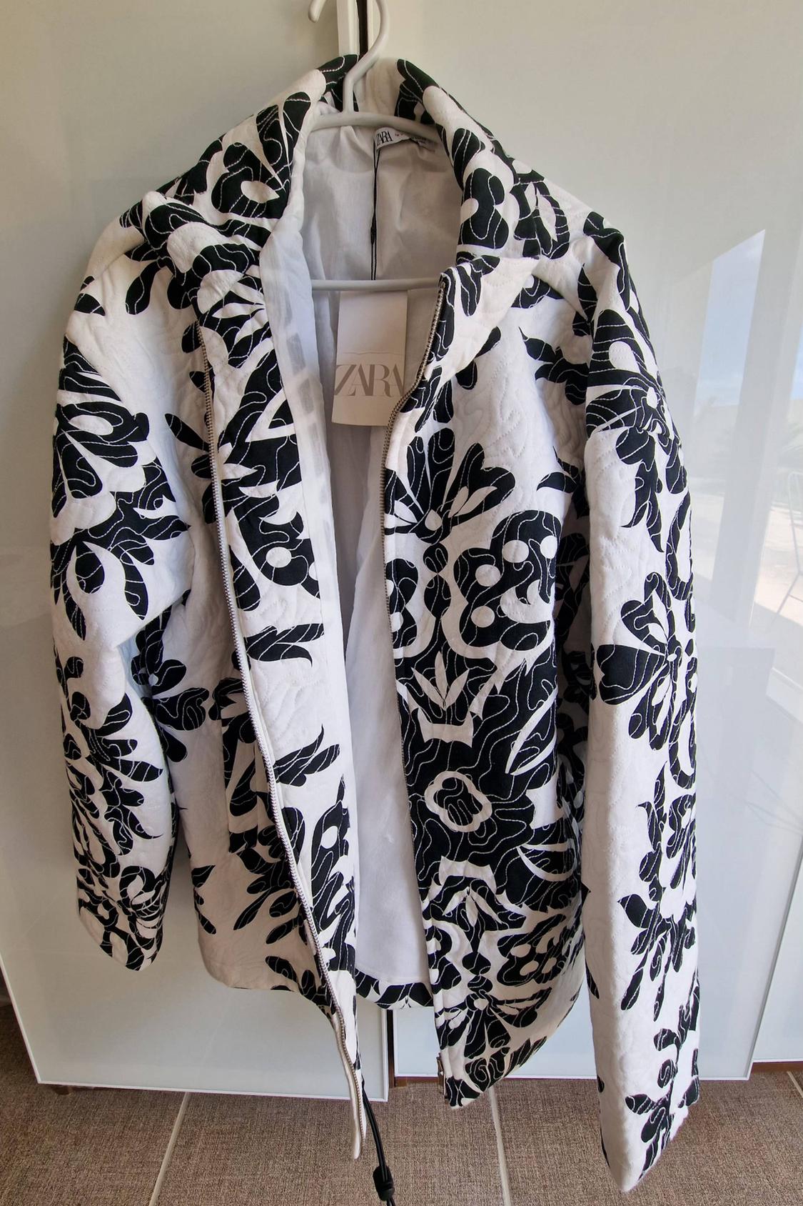 White padded jacket with a black floral print and zip, hanging on a hanger. Label with the text ZARA.