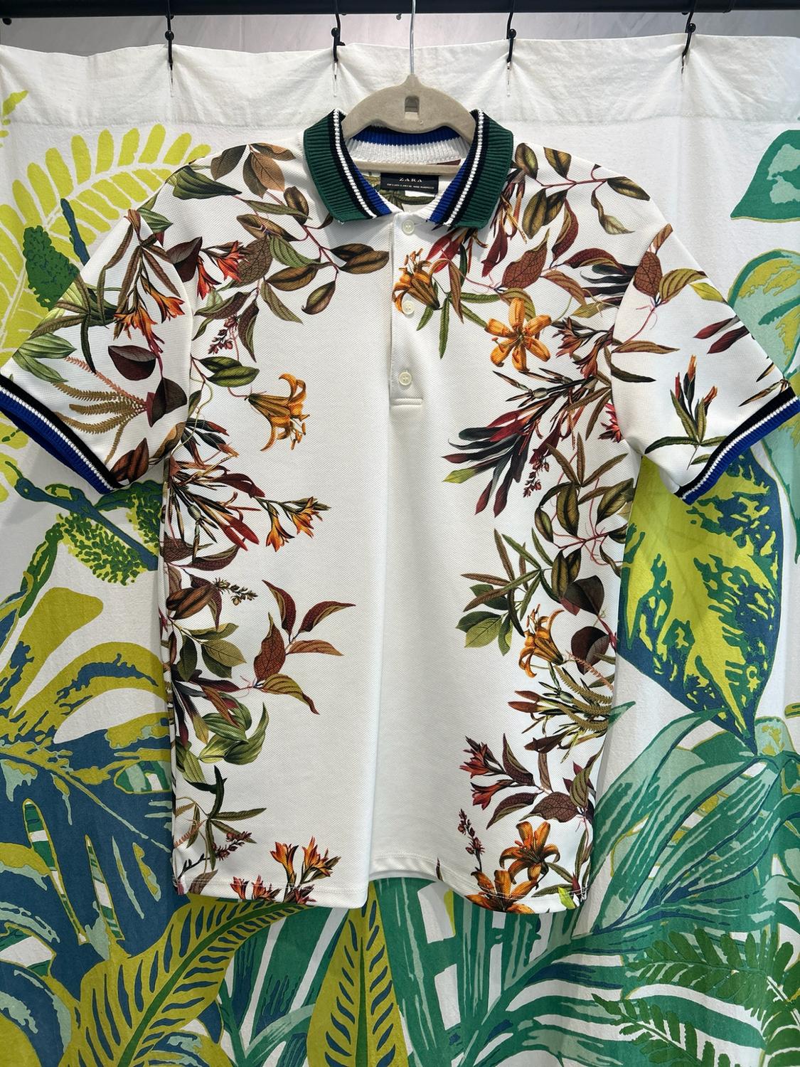 ZARA white polo shirt with a floral print and striped collar, hanging on a hanger against a fabric background with large tropical leaves.