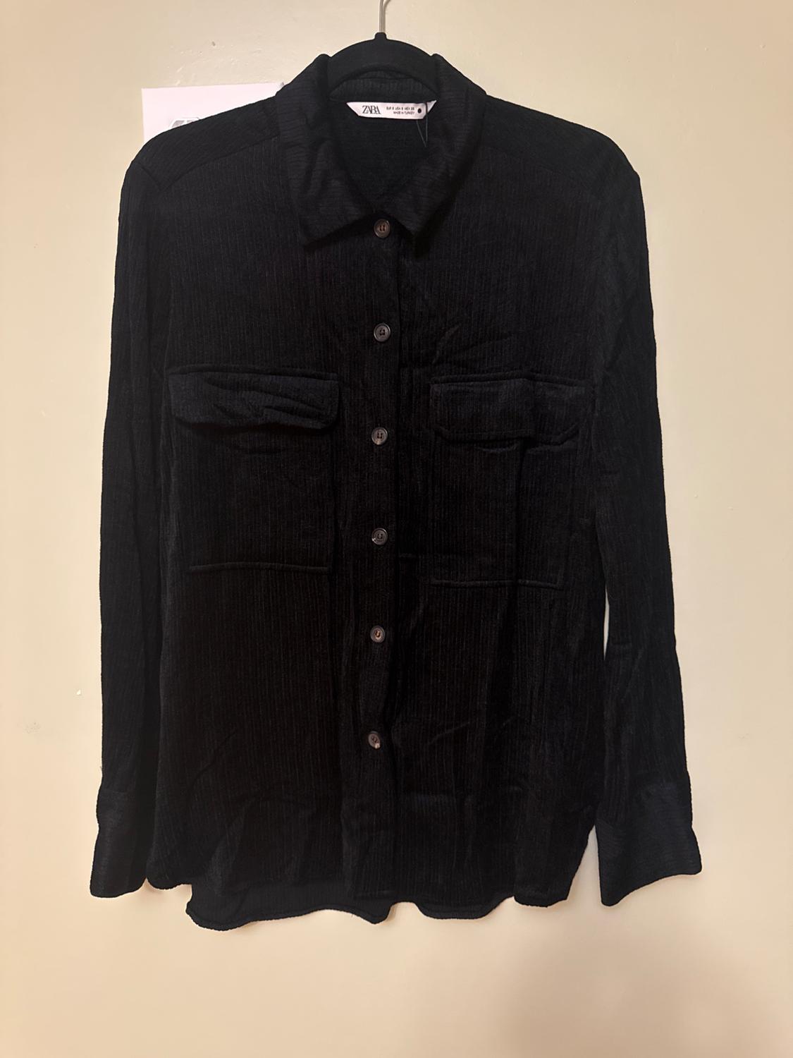 Black corduroy shirt with buttons and two front pockets, hanging on a hanger against a beige background. Visible label on the collar.