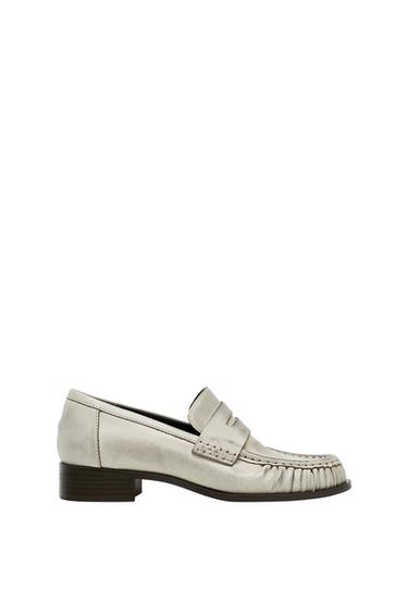 Metallic silver loafer with a mask detail and gathered stitching on the toe. Dark low heel on a white background.