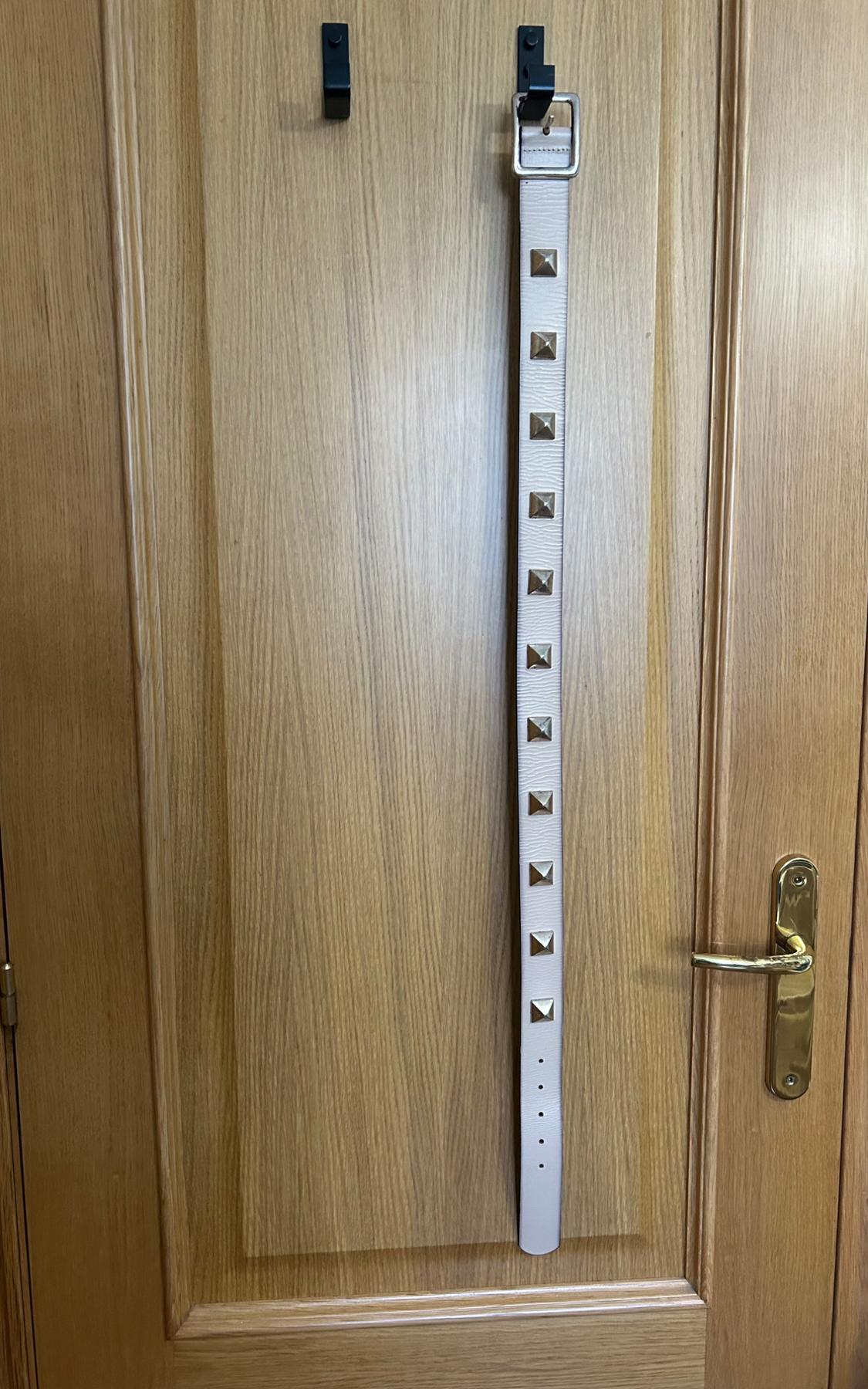 Light-coloured belt with square studs hanging from a black hook on a wooden door with a gold doorknob.