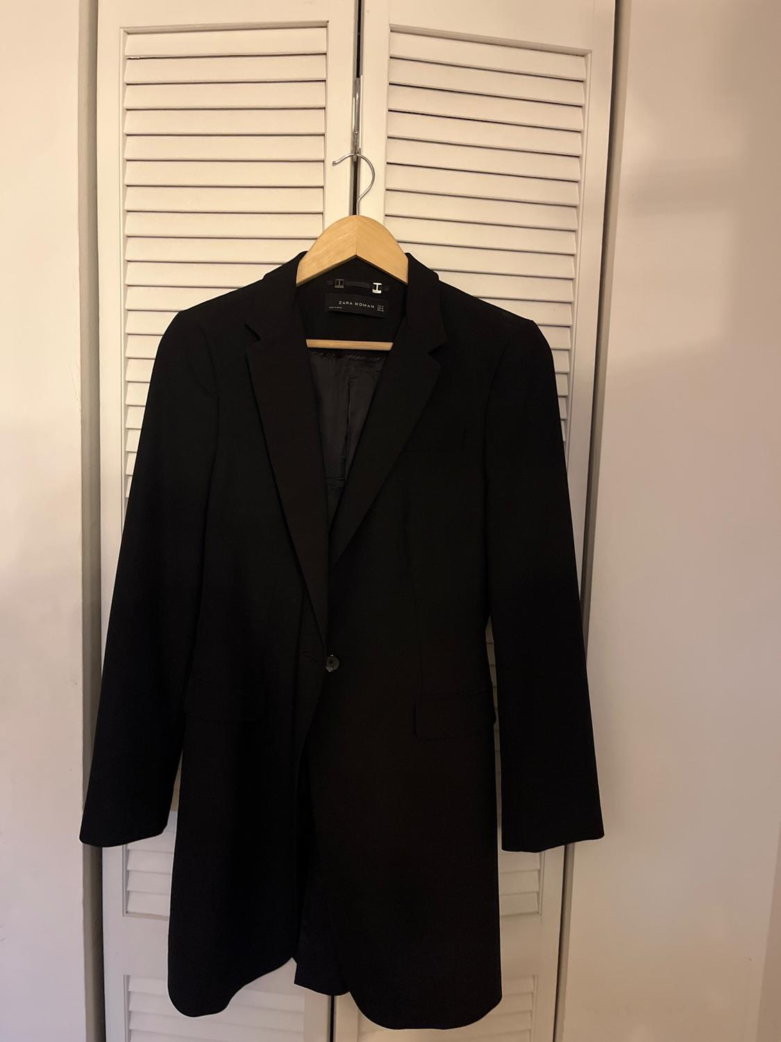 Black jacket hanging on a wooden hanger in front of a white roller door. Visible label: "Zara Woman".