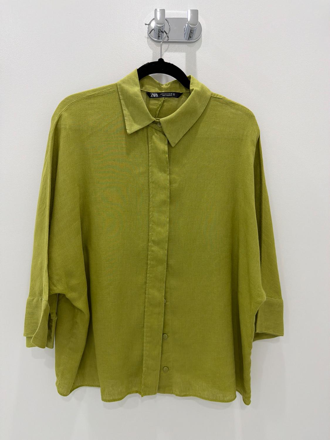 Green button-up shirt hanging on a black hanger against a white wall. Visible text: ZARA.