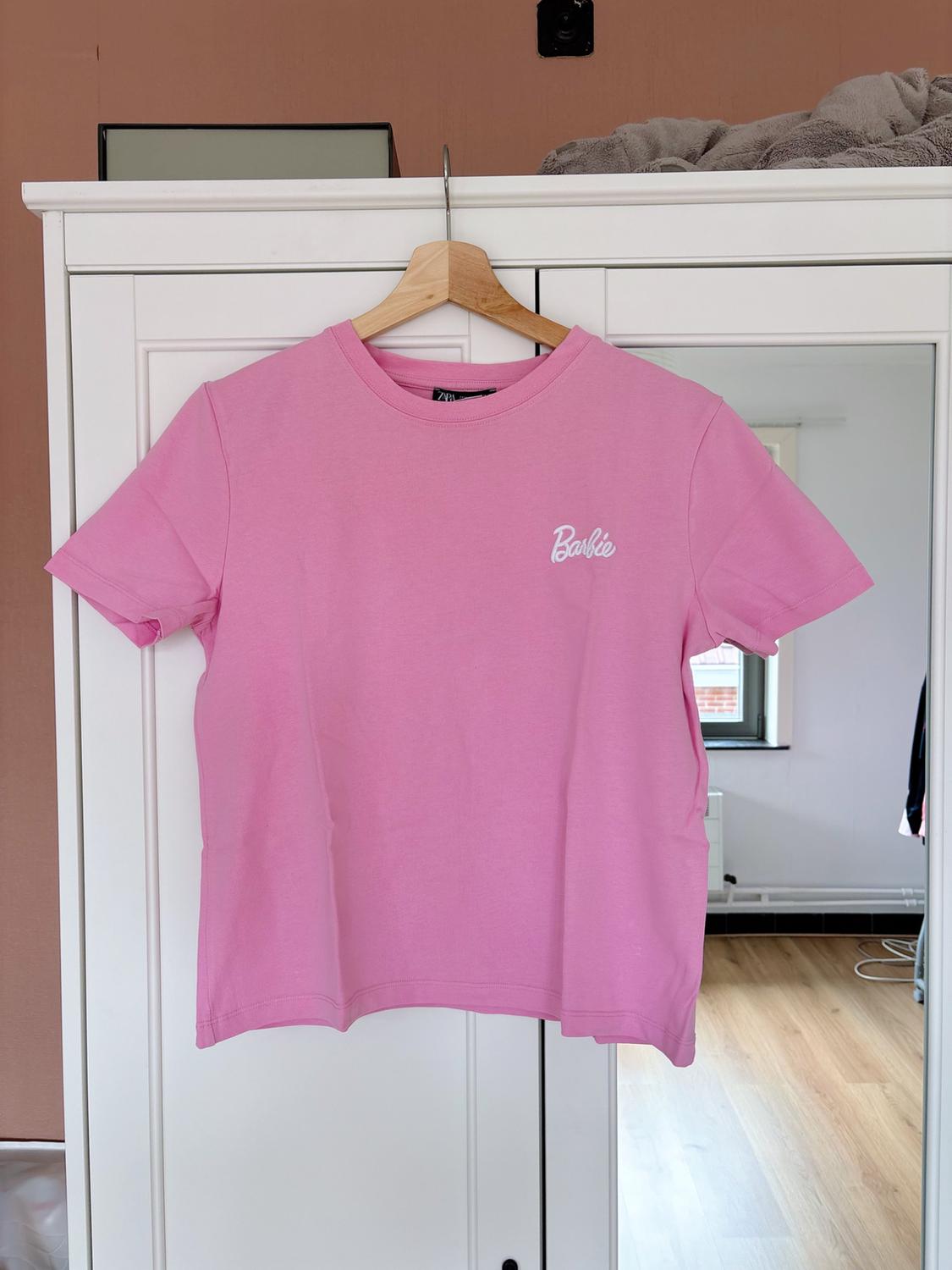 Pink short-sleeved T-shirt with 'Barbie' text, hanging on a wooden hanger in front of a white wardrobe with a mirror.