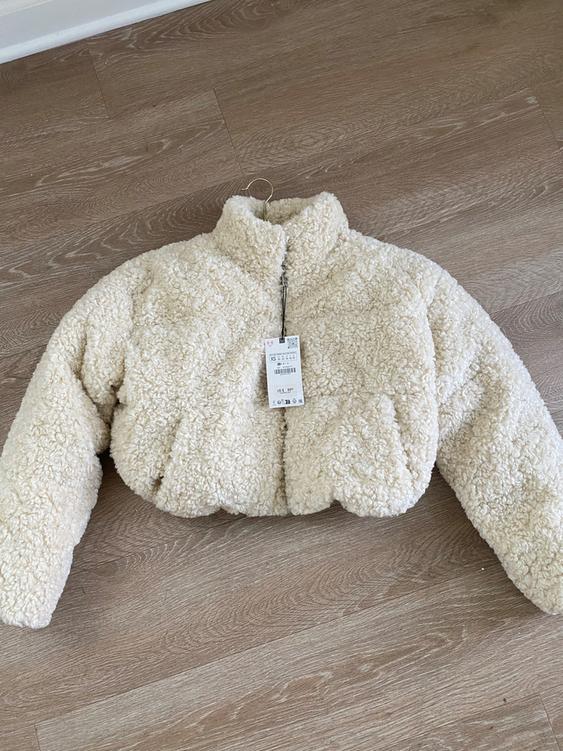 Beige fleece jacket on light wood floor. Visible tag with XS size details and barcode.