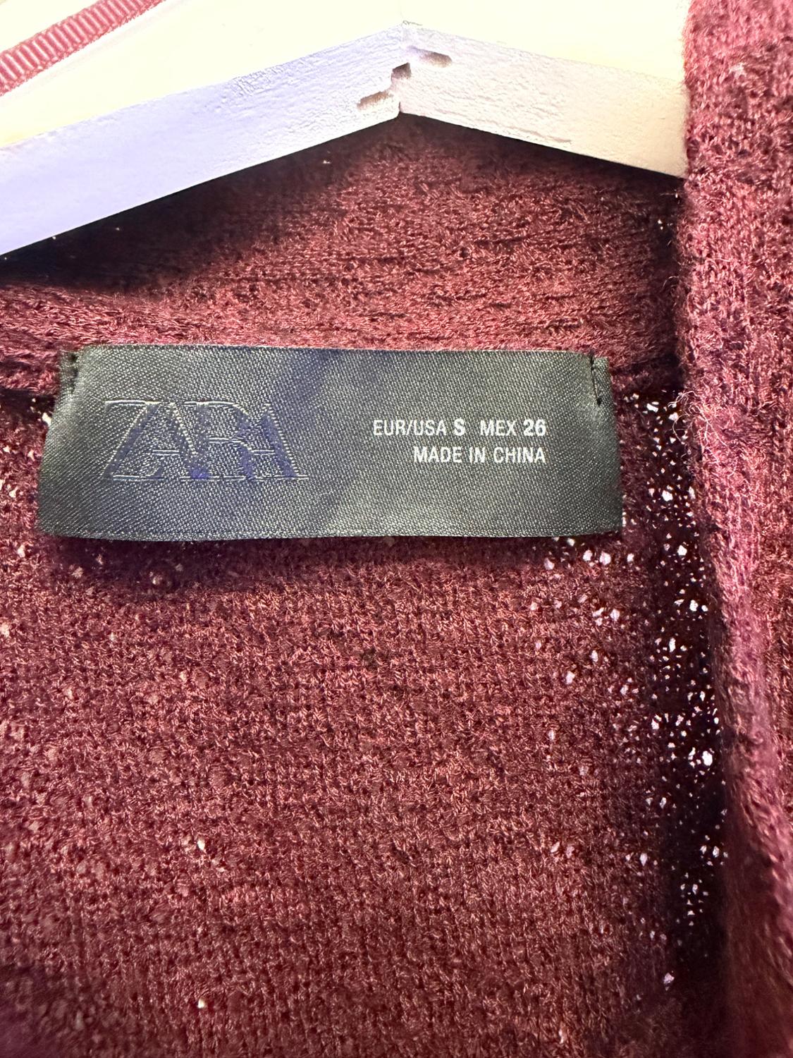 Close-up of a burgundy knitted fabric on a white hanger. Black label with text: ZARA EUR/USA S MEX 26 MADE IN CHINA.