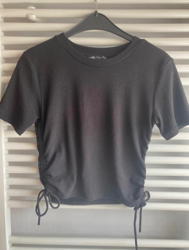 Black short-sleeved t-shirt with side ruching and ties, hanging on a black hanger in front of a white slatted background.