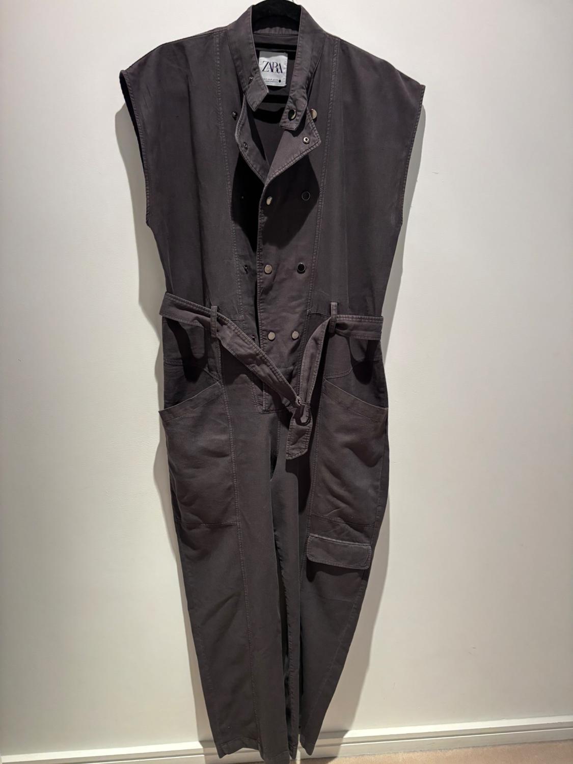 Grey, sleeveless jumpsuit hanging on a hanger, with a belt and large pockets. Visible label with the text "ZARA". White background.