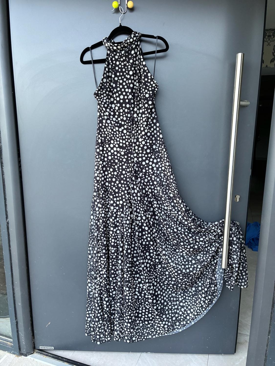 Μάξι μαύρο φόρεμα με λευκά πουά και στράπλες λαιμόκοψη halter, κρεμασμένο σε μια γκρι πόρτα. Στο κάτω μέρος αναγράφεται HORMANN.