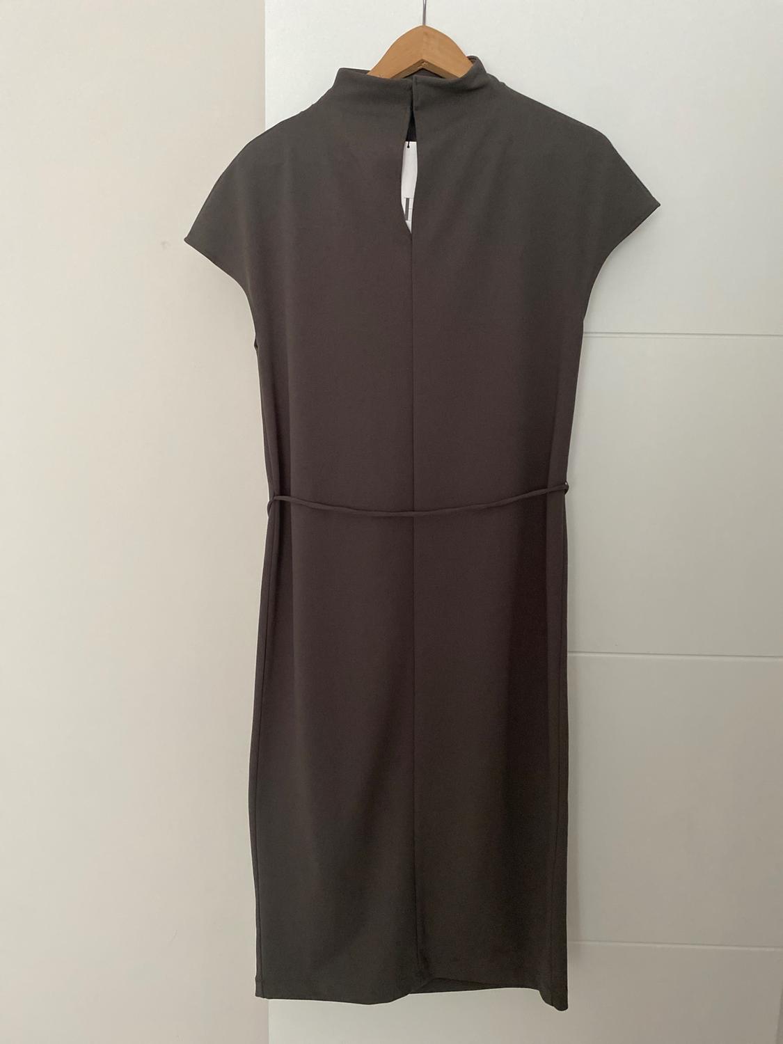 Dark grey sleeveless dress, high neck with a slit and thin belt, hanging on a wooden hanger on a white door. Casual style.