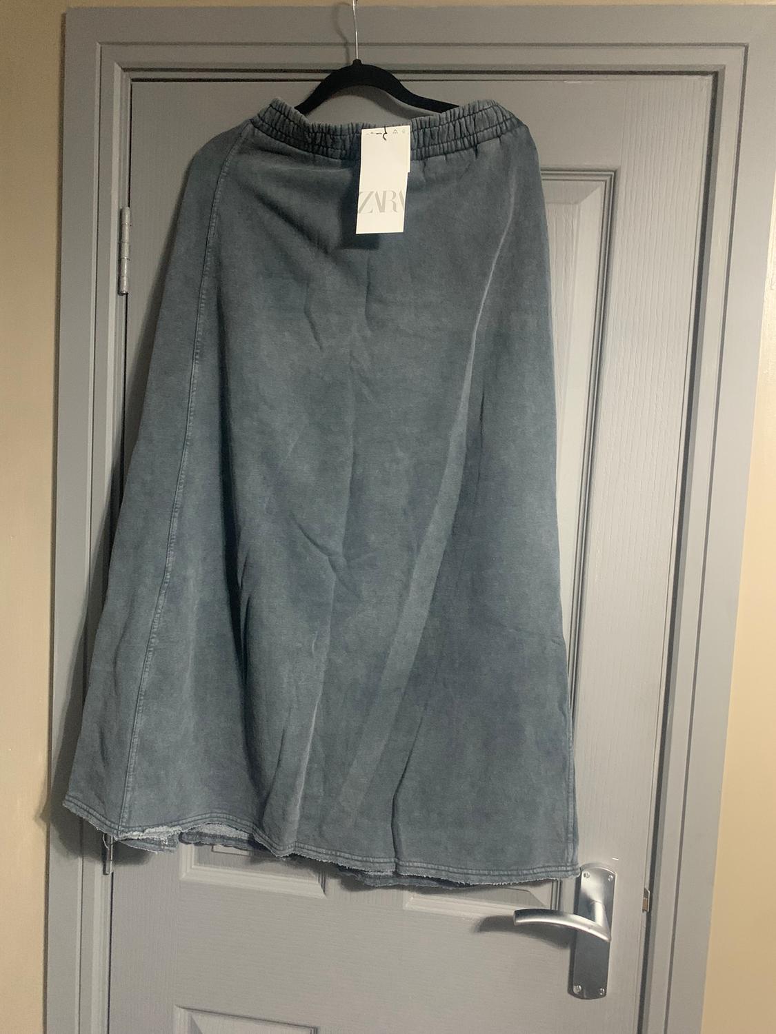 Grey skirt hanging on a door, with a visible label that reads "ZARA". The skirt has an elasticated waist and a simple design.