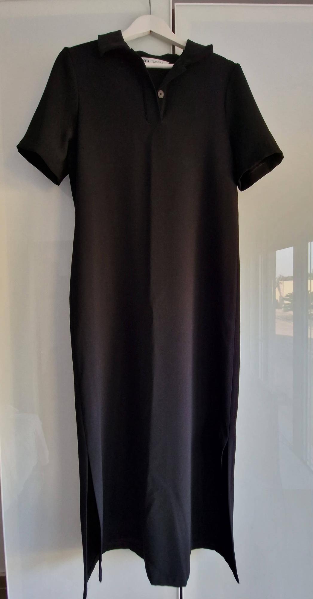 Short-sleeved black dress with a collar, one button and side slits at the hem. It is hanging on a white hanger against a light background.