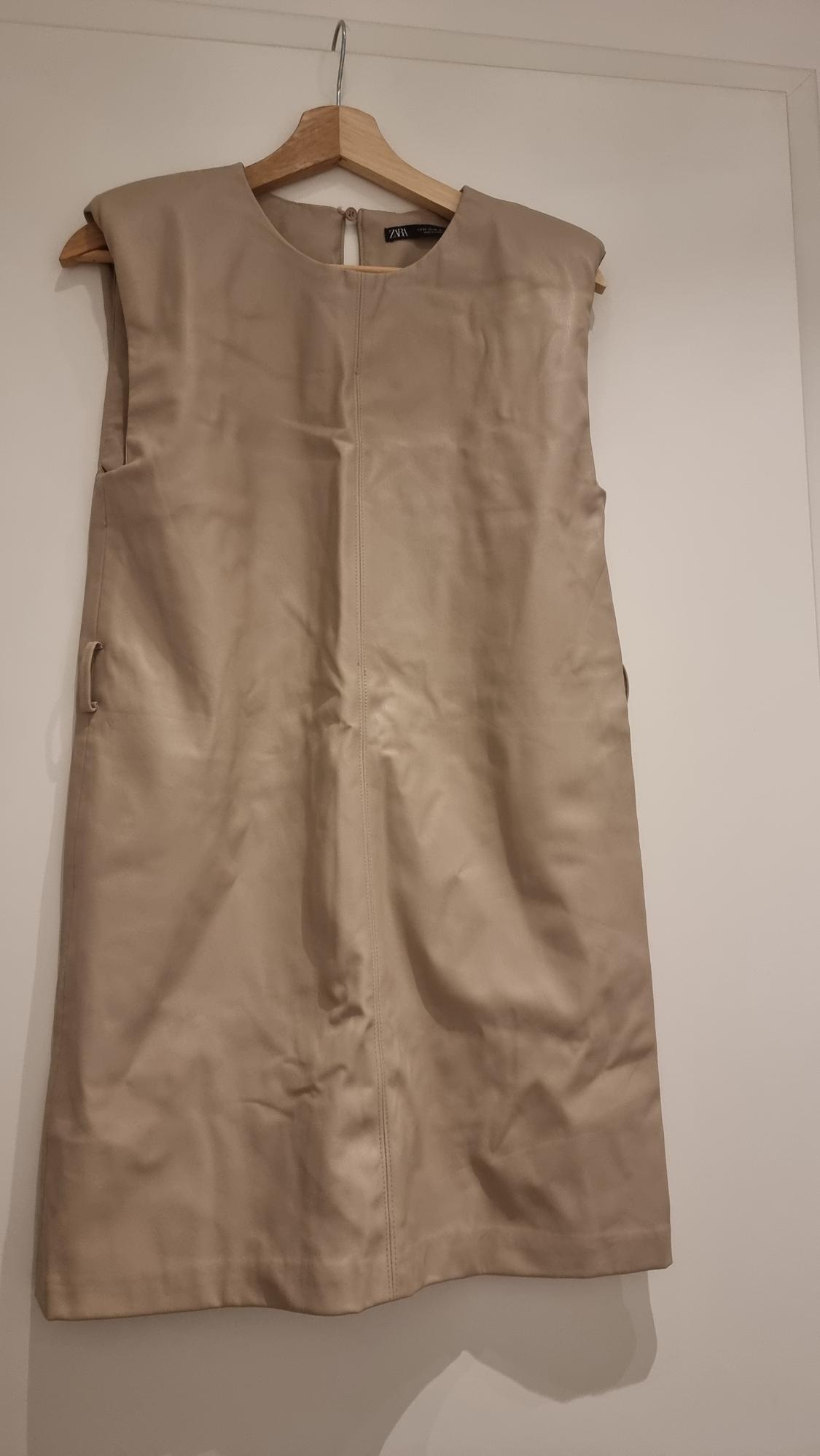 Sleeveless beige dress hanging on a wooden hanger against a white wall. Label visible on the collar.