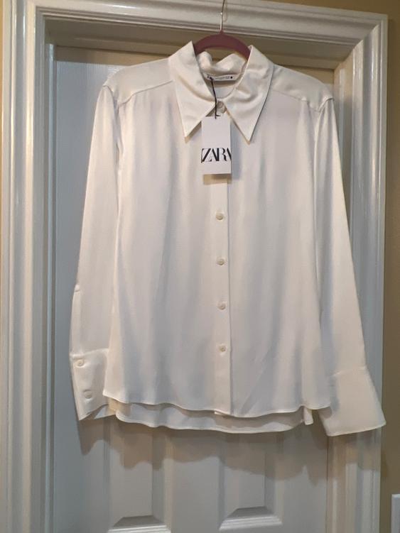 White long-sleeved shirt hanging on a hanger, with a visible tag that says "ZARA", on a white door.