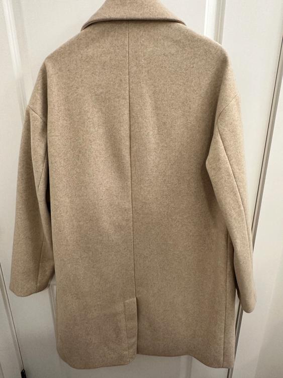 Beige coat hanging on a white door, viewed from behind. Simple design with a classic collar and long sleeves.