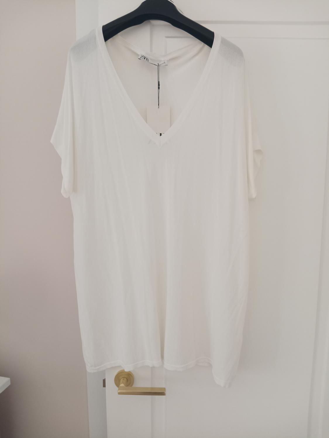 White short-sleeved V-neck T-shirt hanging on a black hanger over a white door. Label visible on the collar.