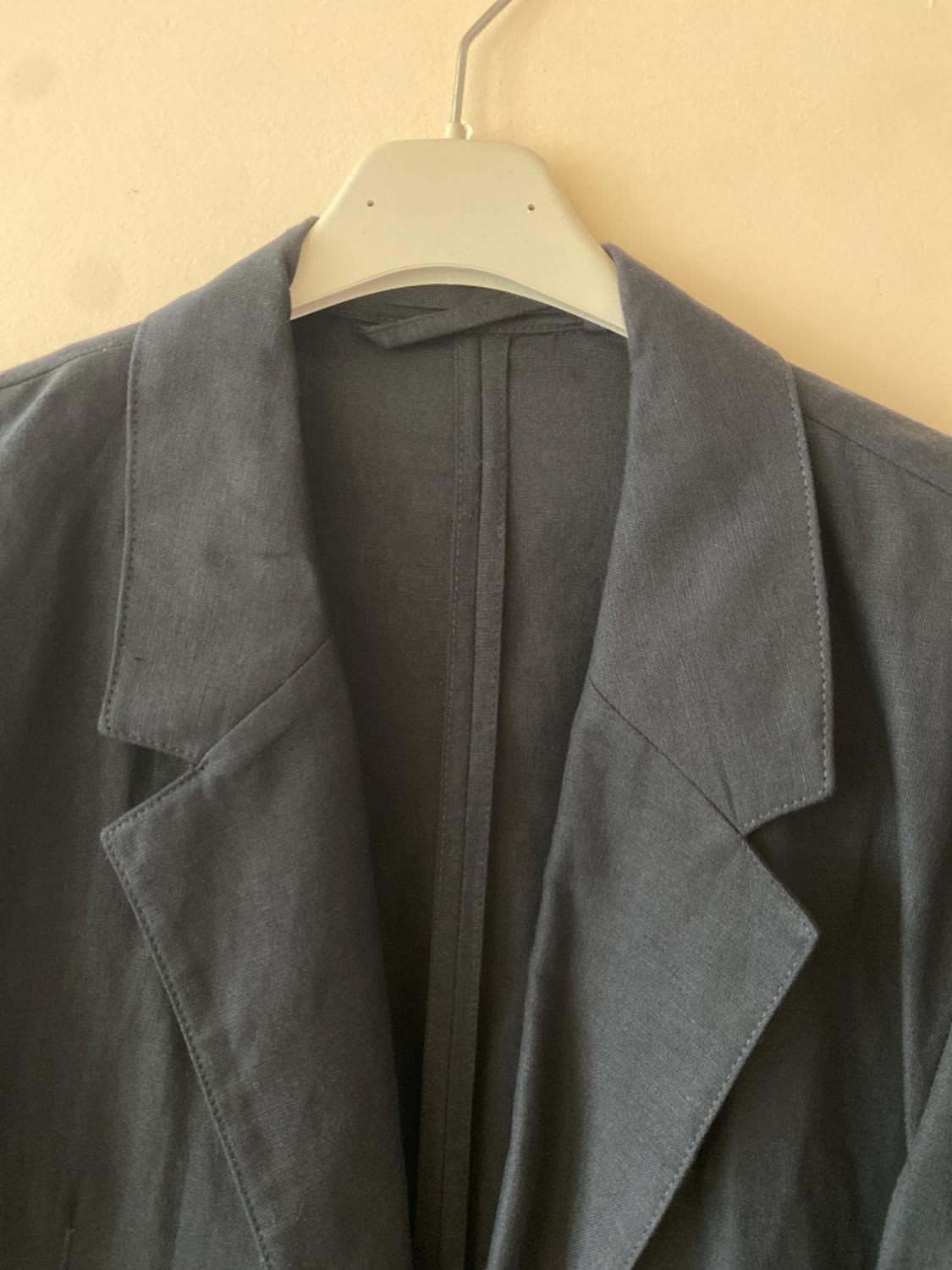 Grey jacket hanging on a white hanger against a beige wall. The jacket has wide lapels and a simple design.