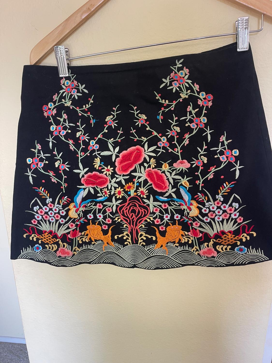 Black skirt with colourful floral embroidery, including red and blue flowers and yellow and orange details, hanging on a hanger.