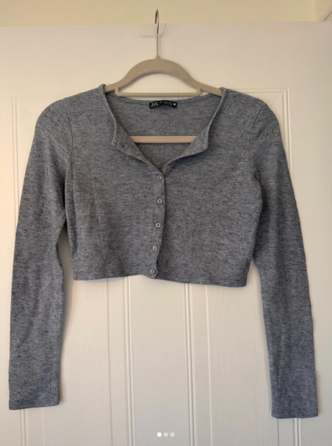 Grey cropped knitted cardigan with long sleeves and buttons, hanging on a hanger on a white door. Inner label with text ZARA.