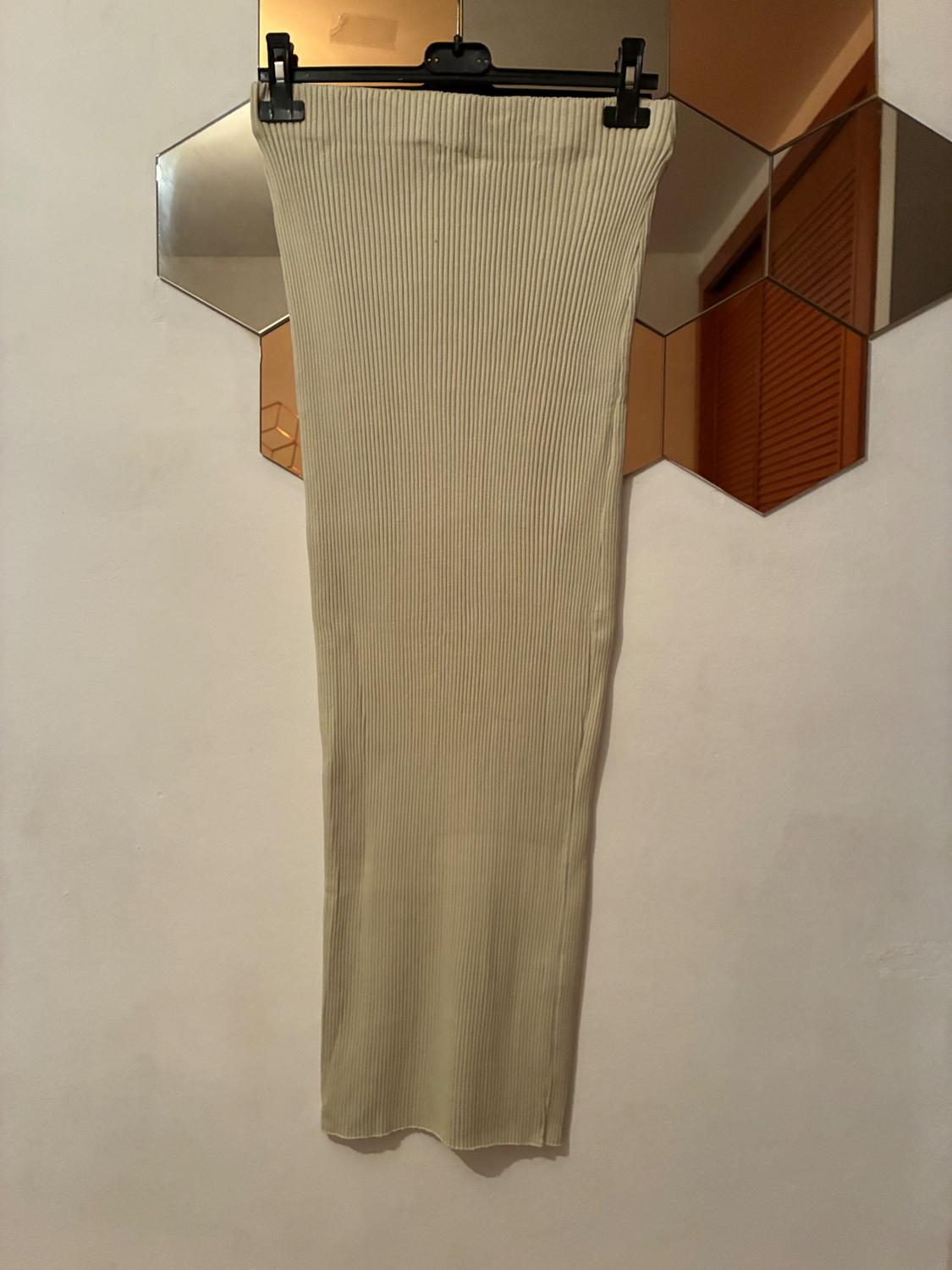 Long ribbed knit skirt in beige, hanging on a black hanger, with a background of hexagonal mirrors on the wall.