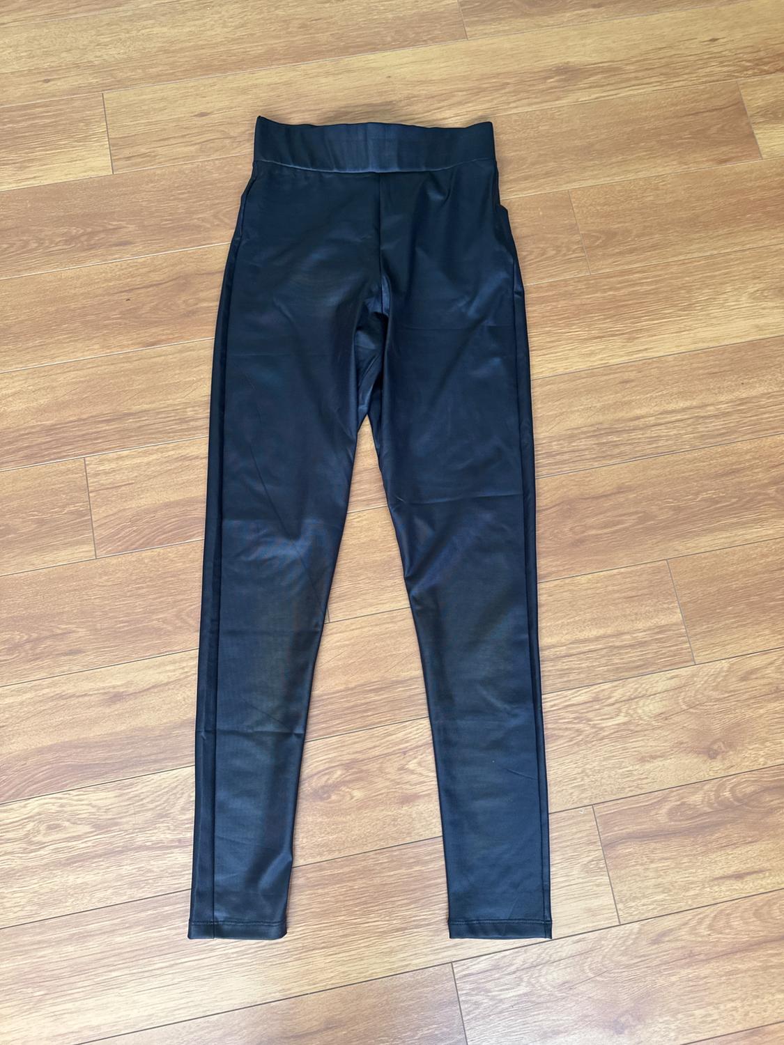 Black trousers made from a shiny material on a light wooden floor. Fitted design with a high waist and clean lines.
