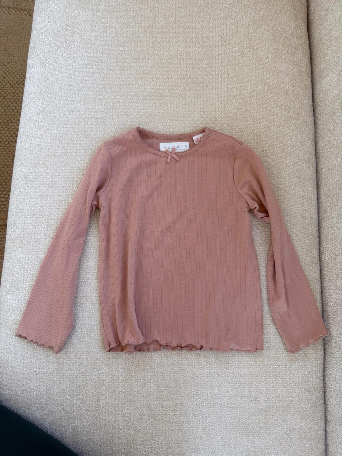 Rosa long-sleeve ribbed T-shirt with a bow at the neckline and wavy edges, on a beige sofa. Text: ZARA 5 110.