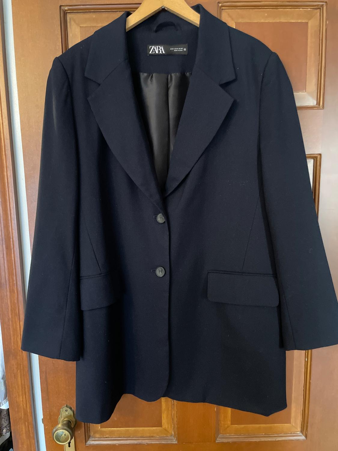 Women's black jacket hanging on a wooden hanger. Visible "ZARA" brand tag. Wooden door in the background.