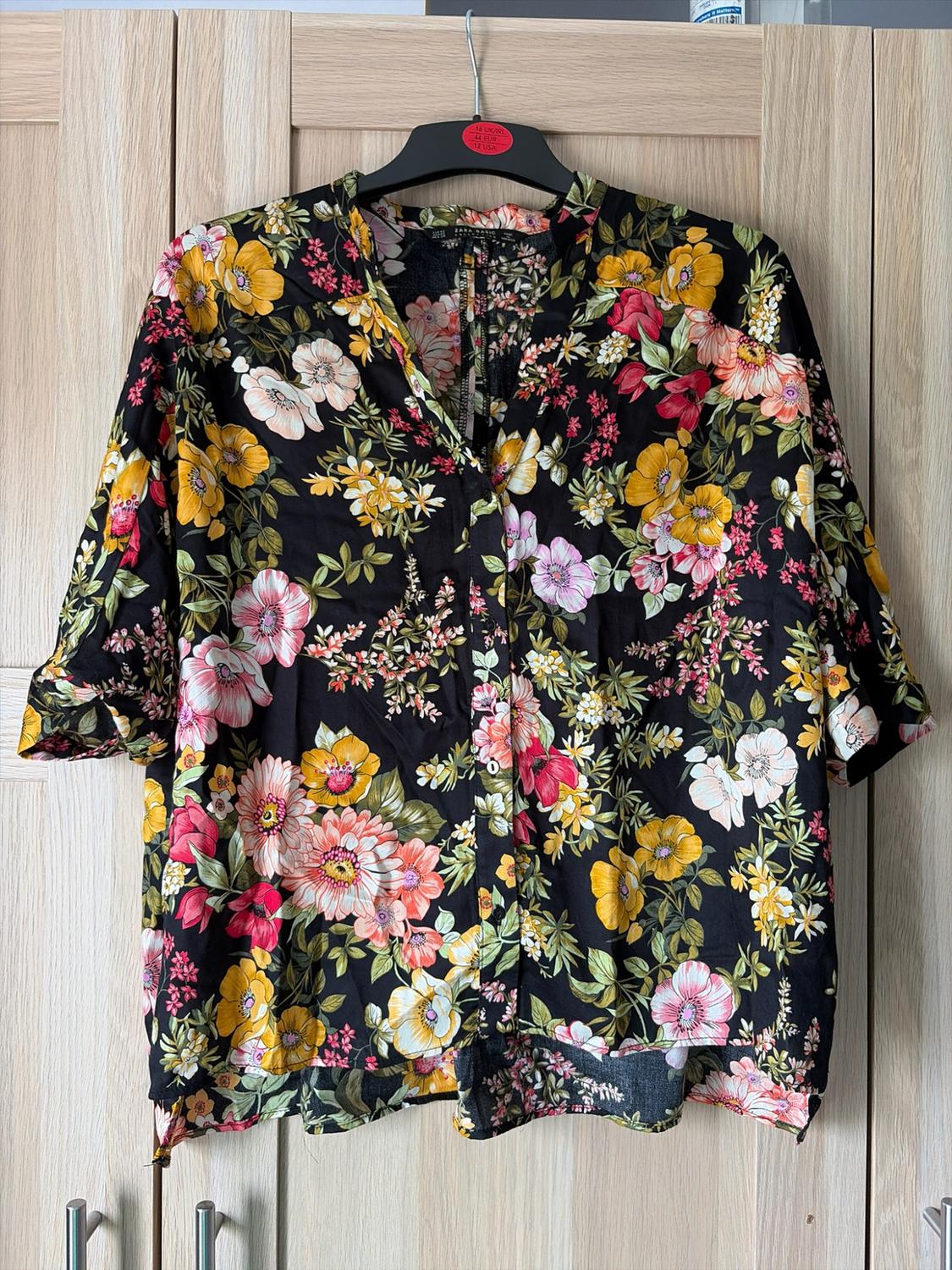 Black shirt with pink and yellow floral print hanging in a wooden wardrobe. Text: 16 UK/IRL 44 EUR 12 USA ZARA BASIC.