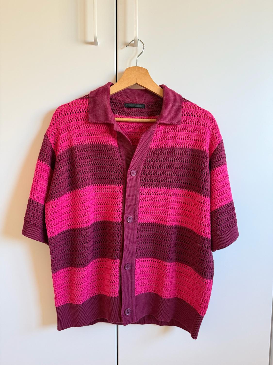 Open-knit, short-sleeved shirt with horizontal pink and burgundy stripes, hanging on a wooden hanger in front of a white wardrobe.