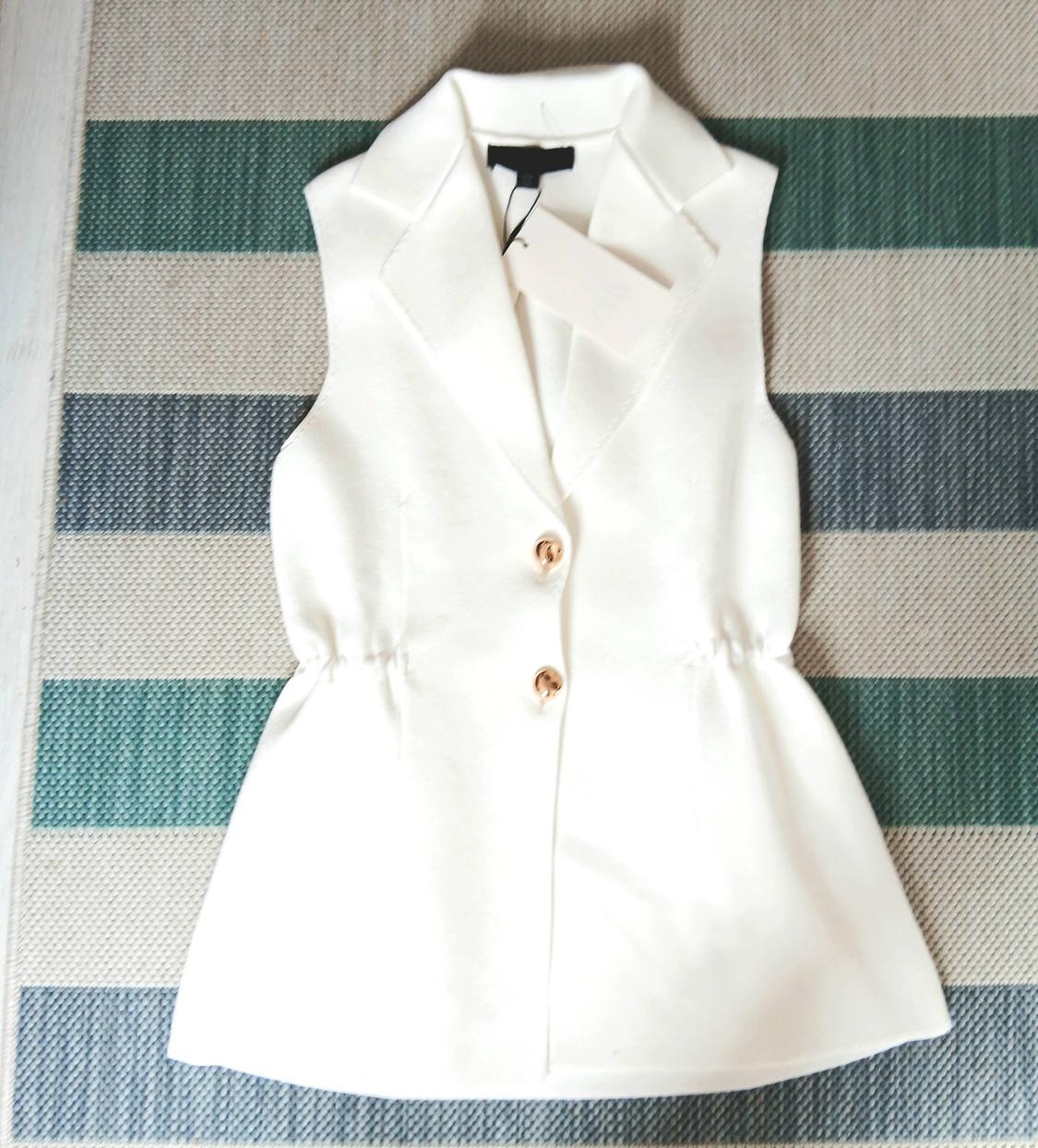 White sleeveless waistcoat with brown buttons on a green and grey striped rug. Hang tag visible.