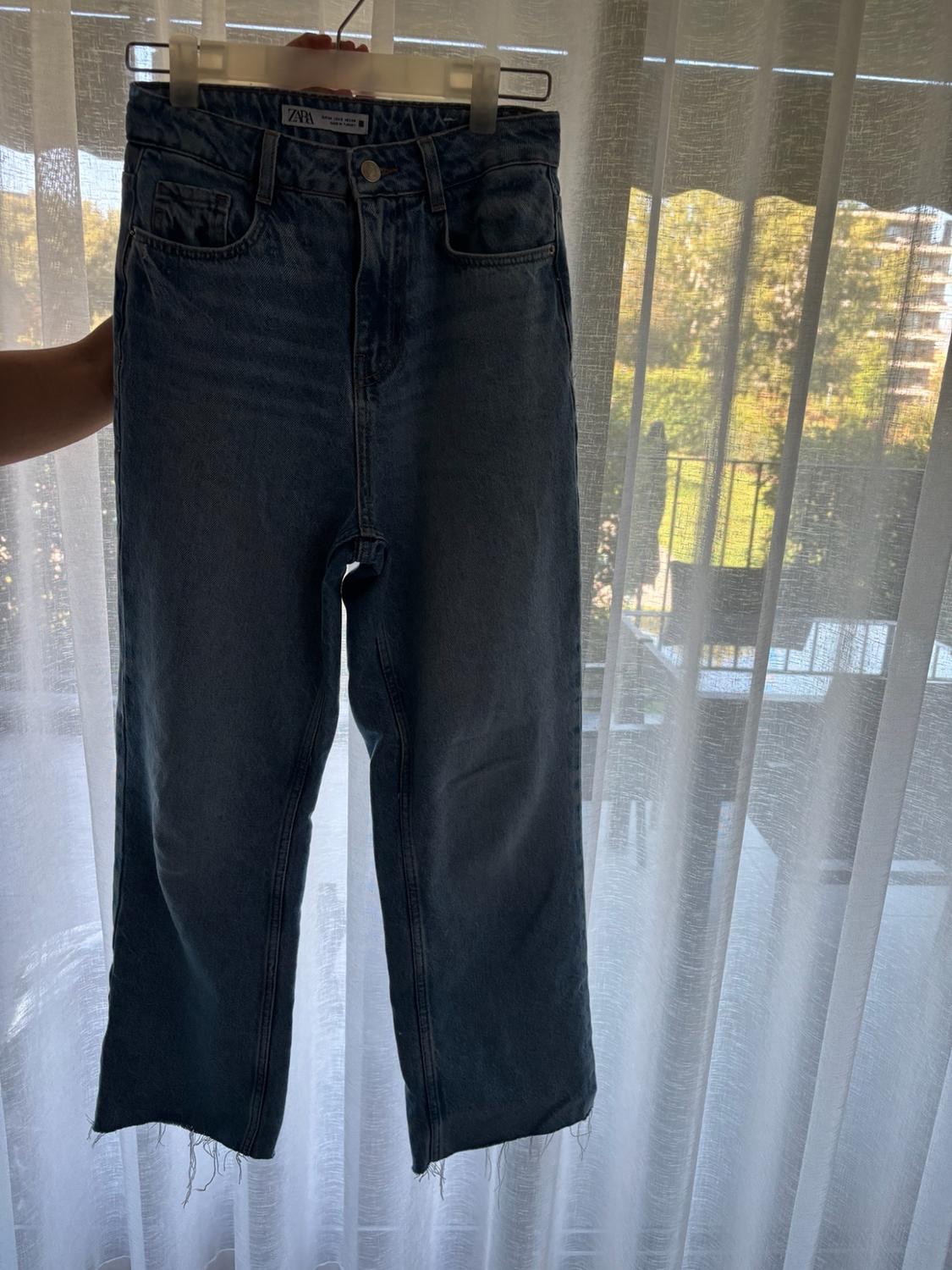 Blue ZARA jeans with a frayed hem on a hanger, held up in front of a window with white curtains and natural light.