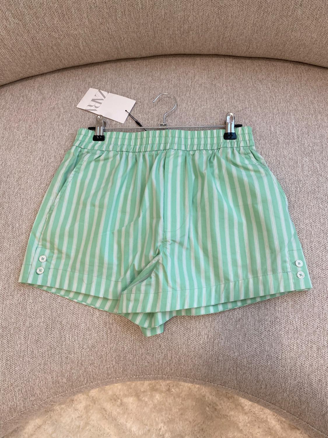 Green shorts with white stripes hanging on a hook above a beige sofa. Visible tag with text "ZARA".