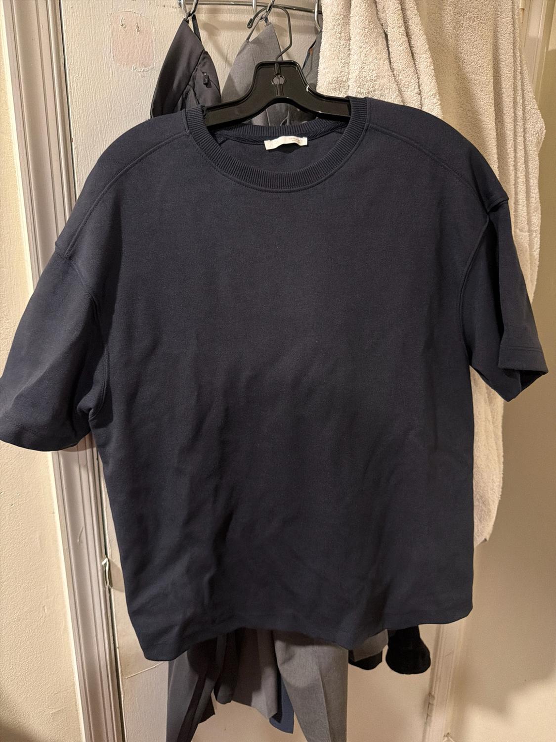 Short-sleeved navy blue T-shirt hanging on a black hanger on a door, with other garments and a towel in the background. Casual style.