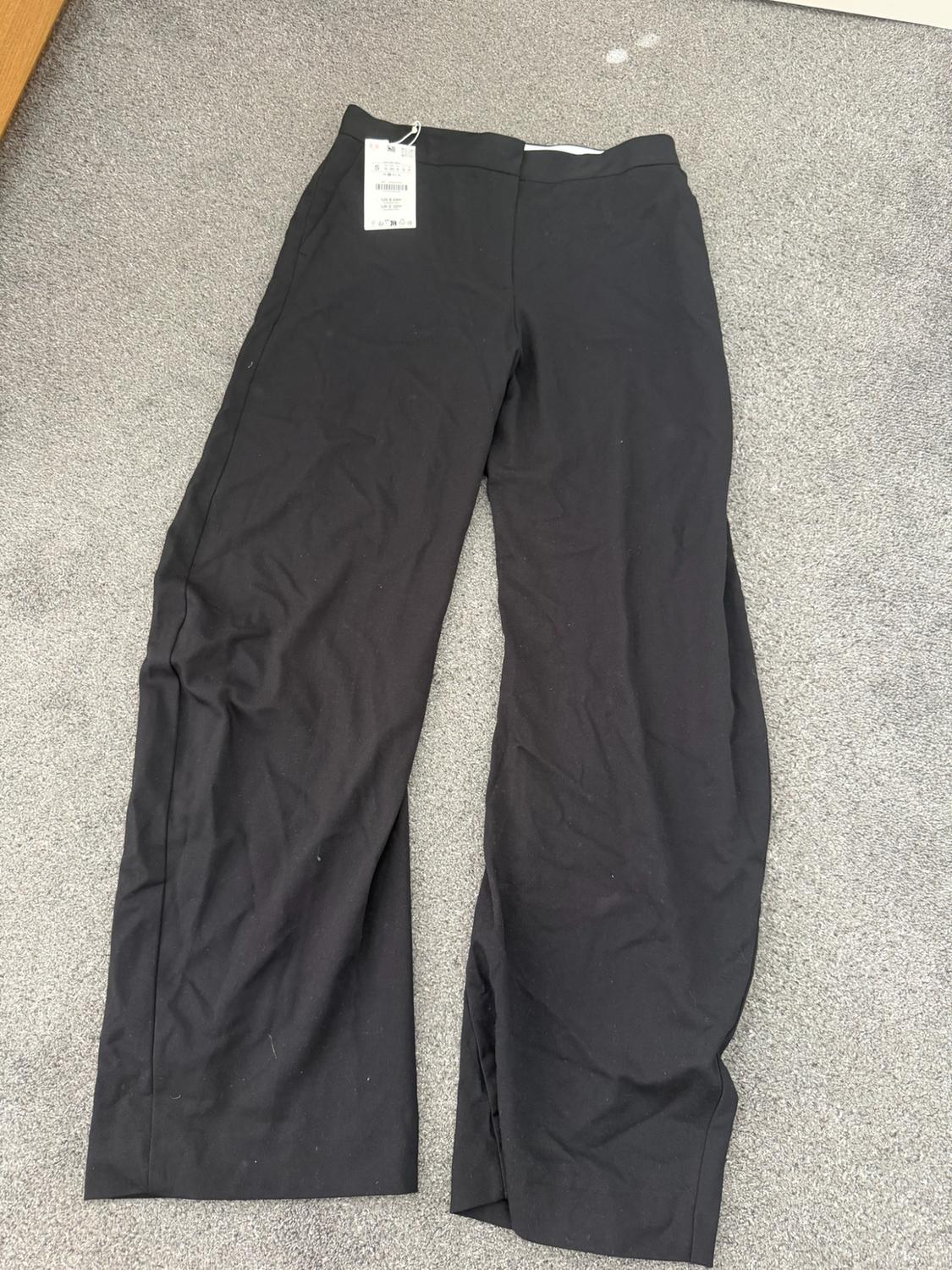 Black wide-leg trousers with a label, laid out on a grey carpet.