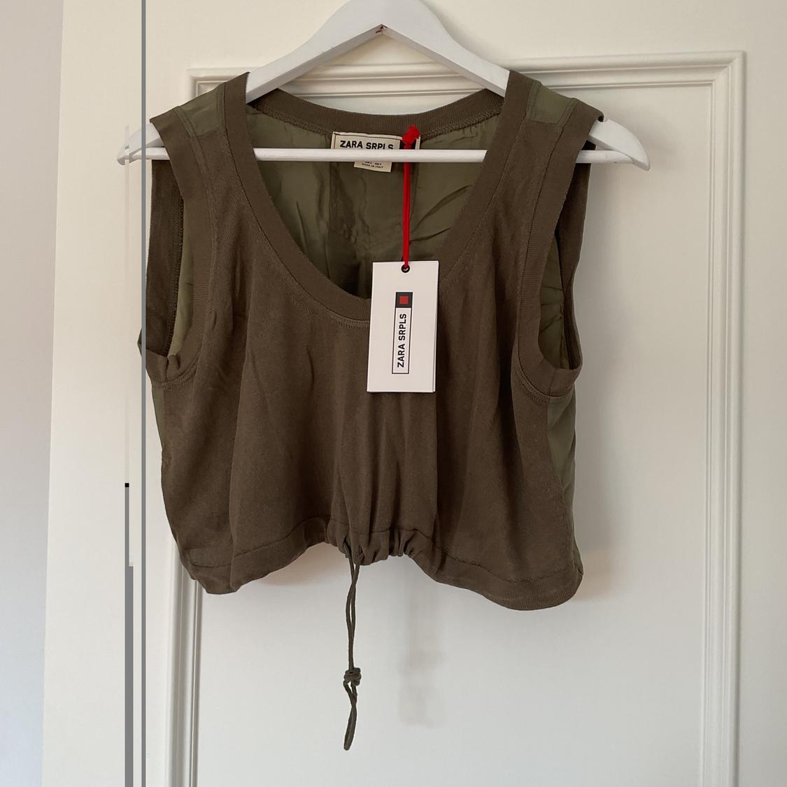 Khaki sleeveless top hanging on a white hanger, with a "ZARA SRPLS" tag hanging from a red cord.
