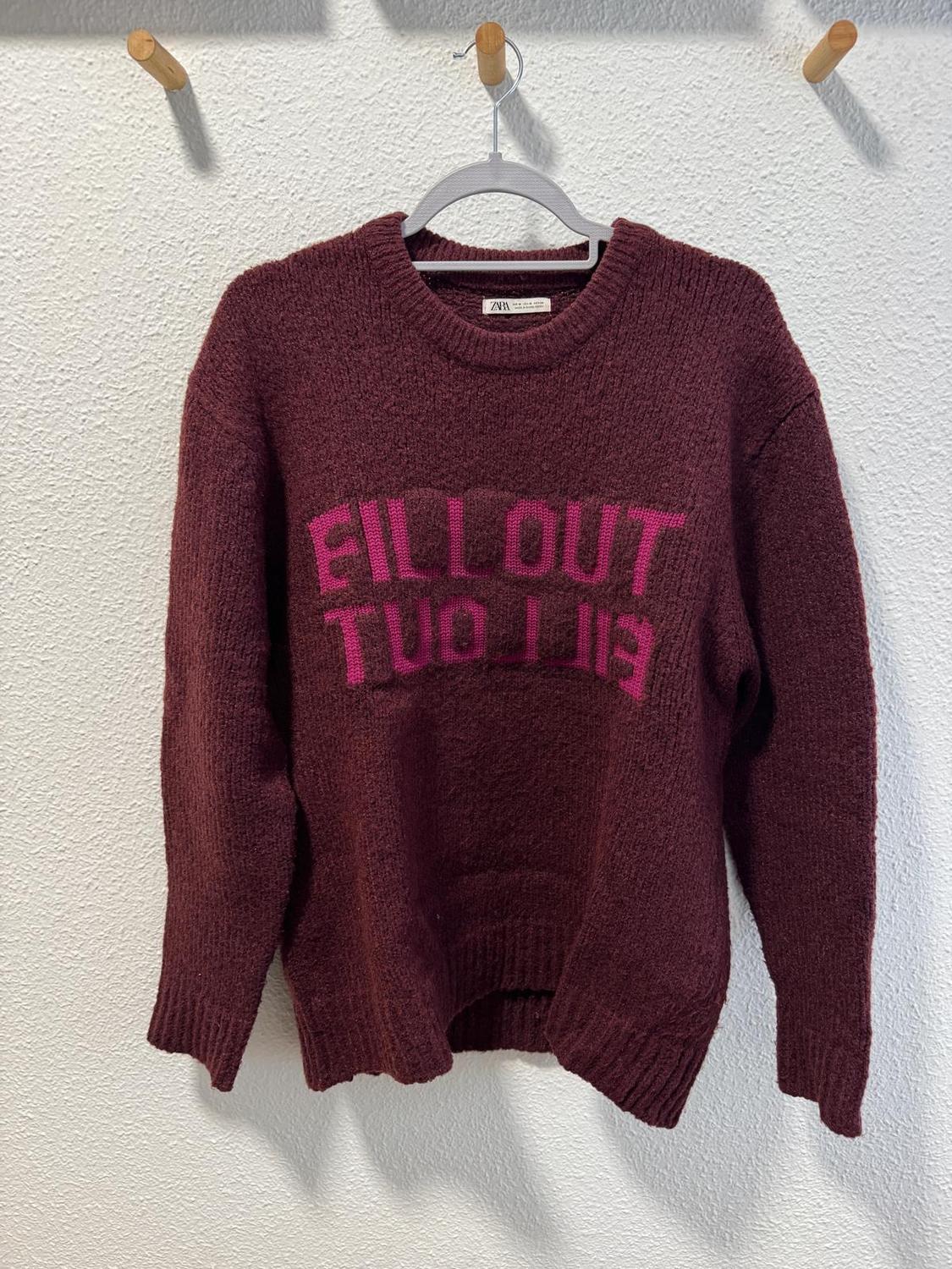 Burgundy jumper with pink text "FILL OUT TWO LINES" hanging on a hanger against a white background.