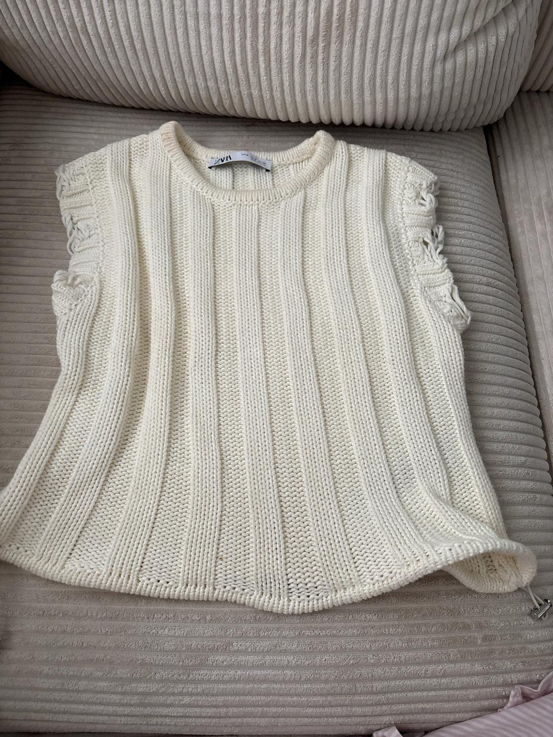 Cream sleeveless knitted top with a ribbed design and details around the armholes, on a corduroy sofa. Visible text: ZARA.