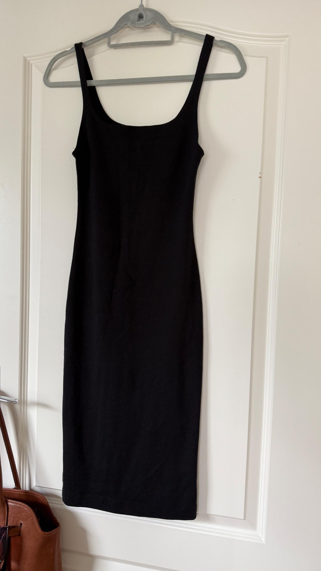 Black sleeveless dress hanging on a grey hanger on a white door. On the left, a brown bag is partially visible.