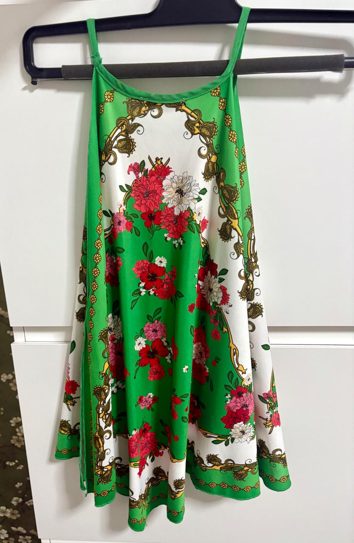 Dress hanging on a hanger, with a floral pattern in green, red, and white tones, and gold details. White and green background.