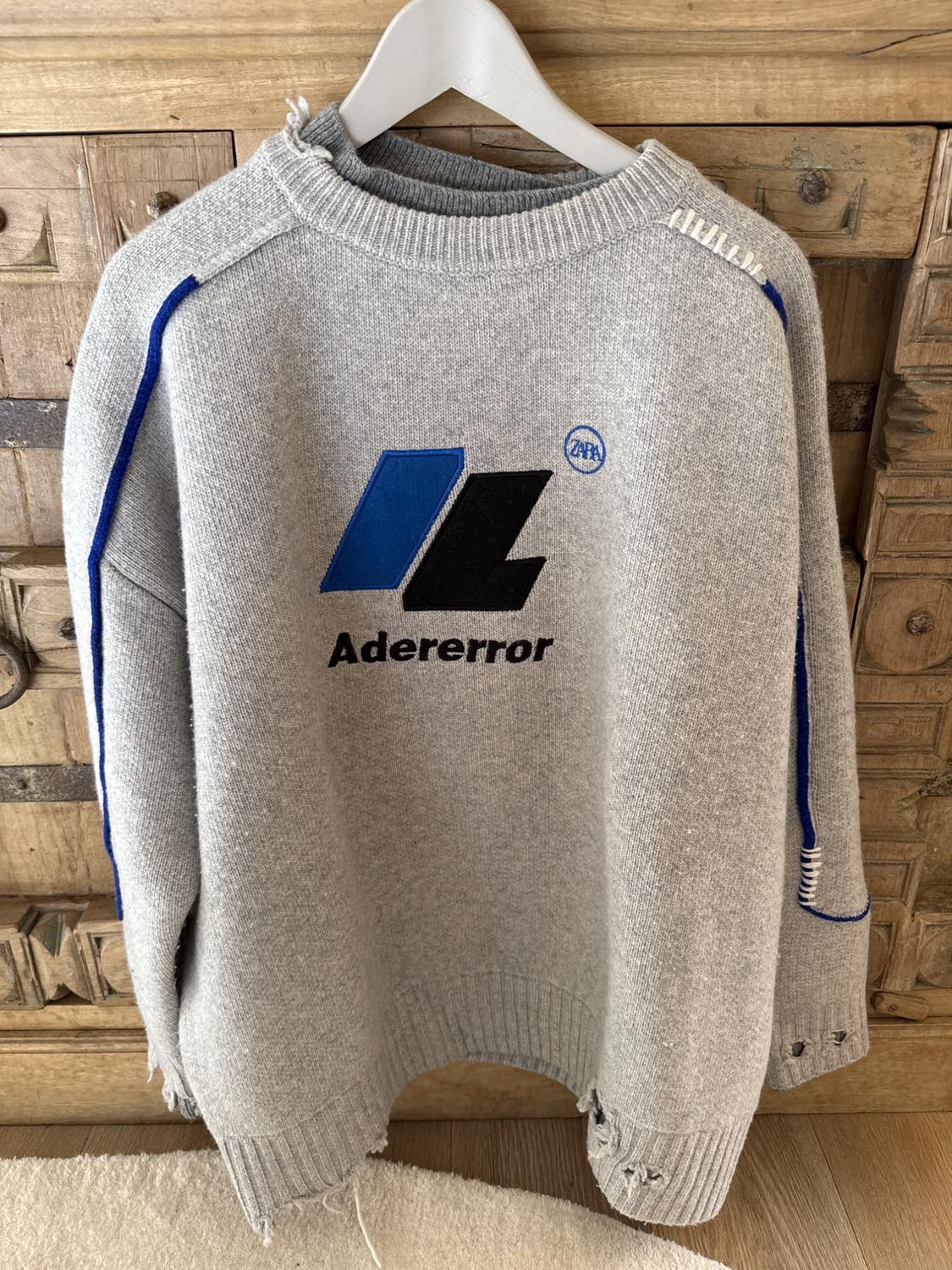 Grey jumper with blue and black details, "Adererror" text on the front, hanging on a white hanger against a wooden background.