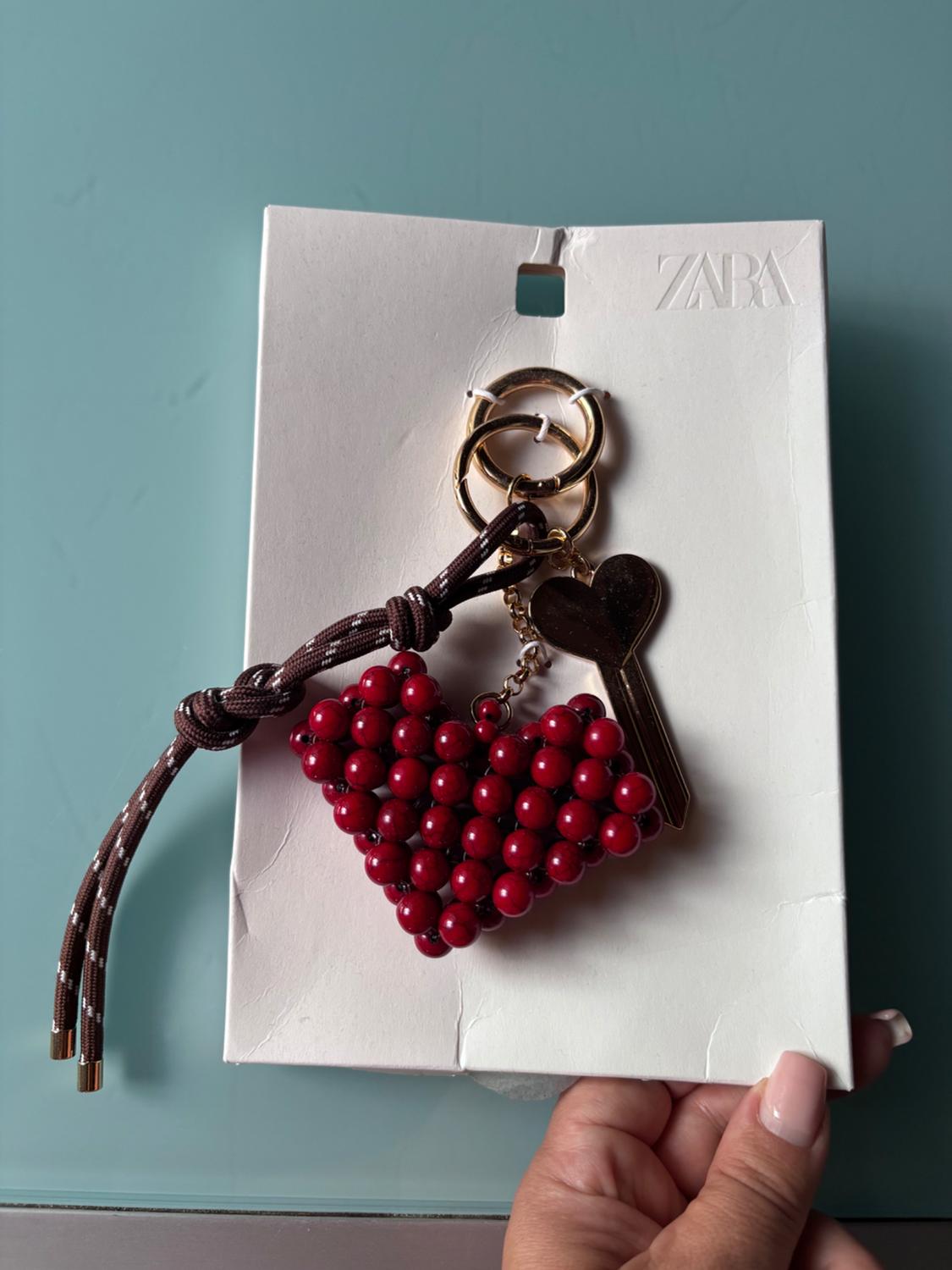 Hand holding a white tag with the text ZARA. Keyring with a red-beaded heart, a gold key and a brown cord against a blue background.