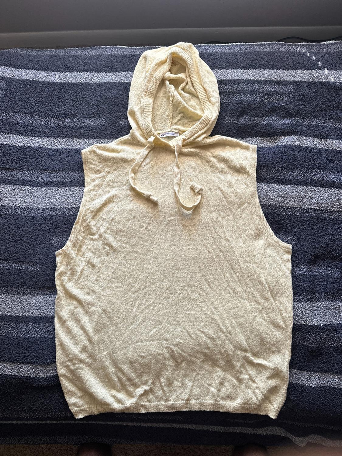 Light yellow knit hooded sleeveless sweatshirt with drawstrings, spread out on a blue and gray striped blanket.